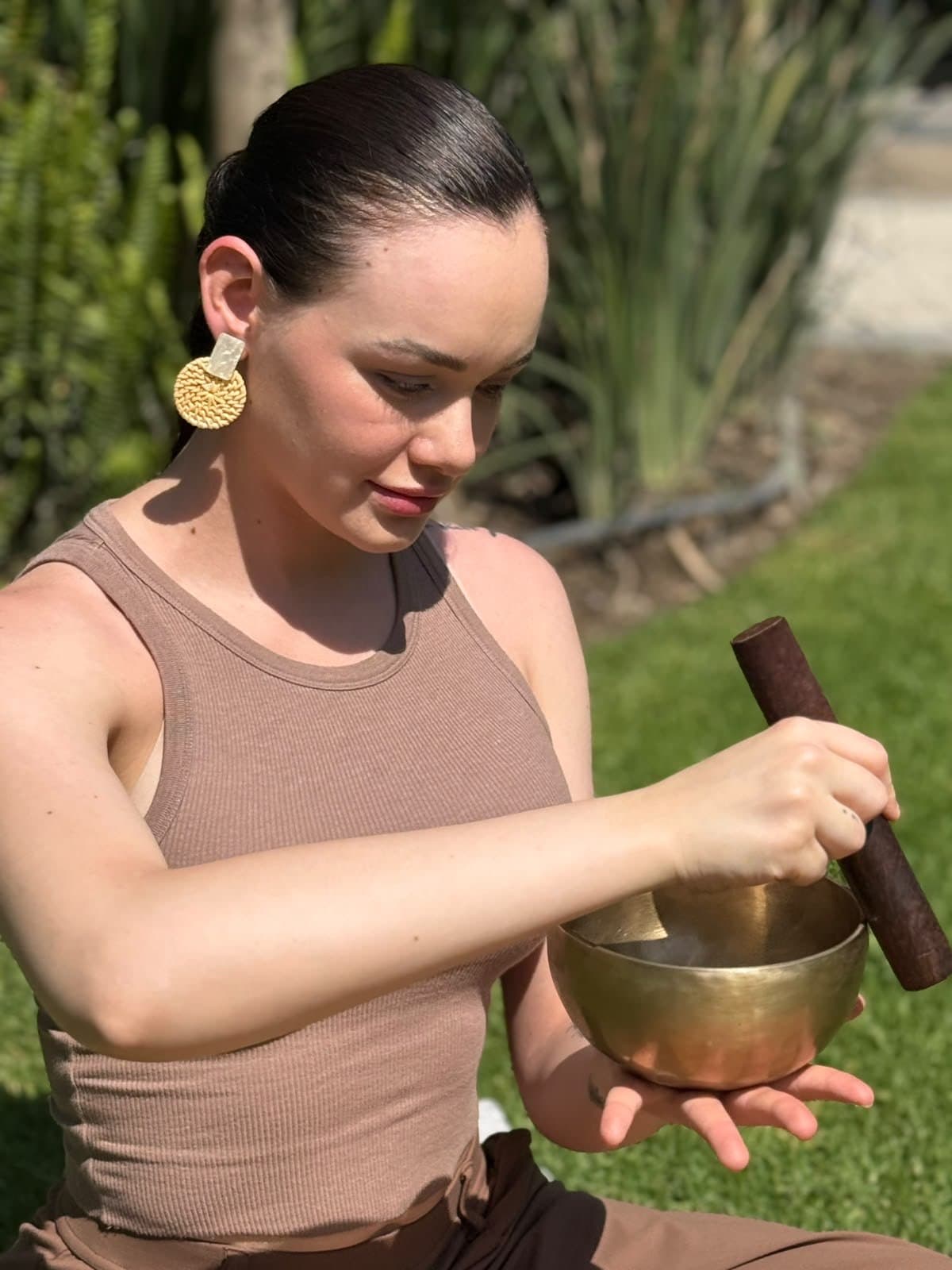 Erika, sentada al aire libre, sosteniendo un cuenco de metal y recorriendo su borde con un mazo de madera.