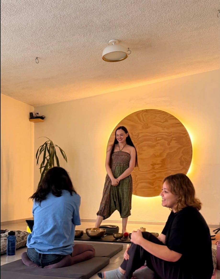 Erika de pie frente a un círculo de madera iluminado, con sus cuencos en el piso y dos personas sentadas en tapetes.